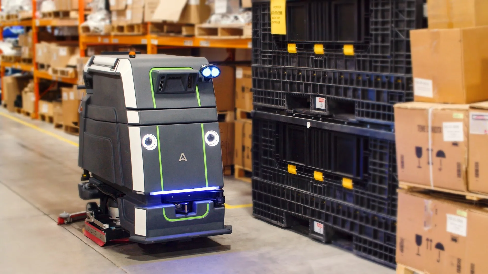 An autonomous floor scrubber working a warehouse aisle beside stacked pallets